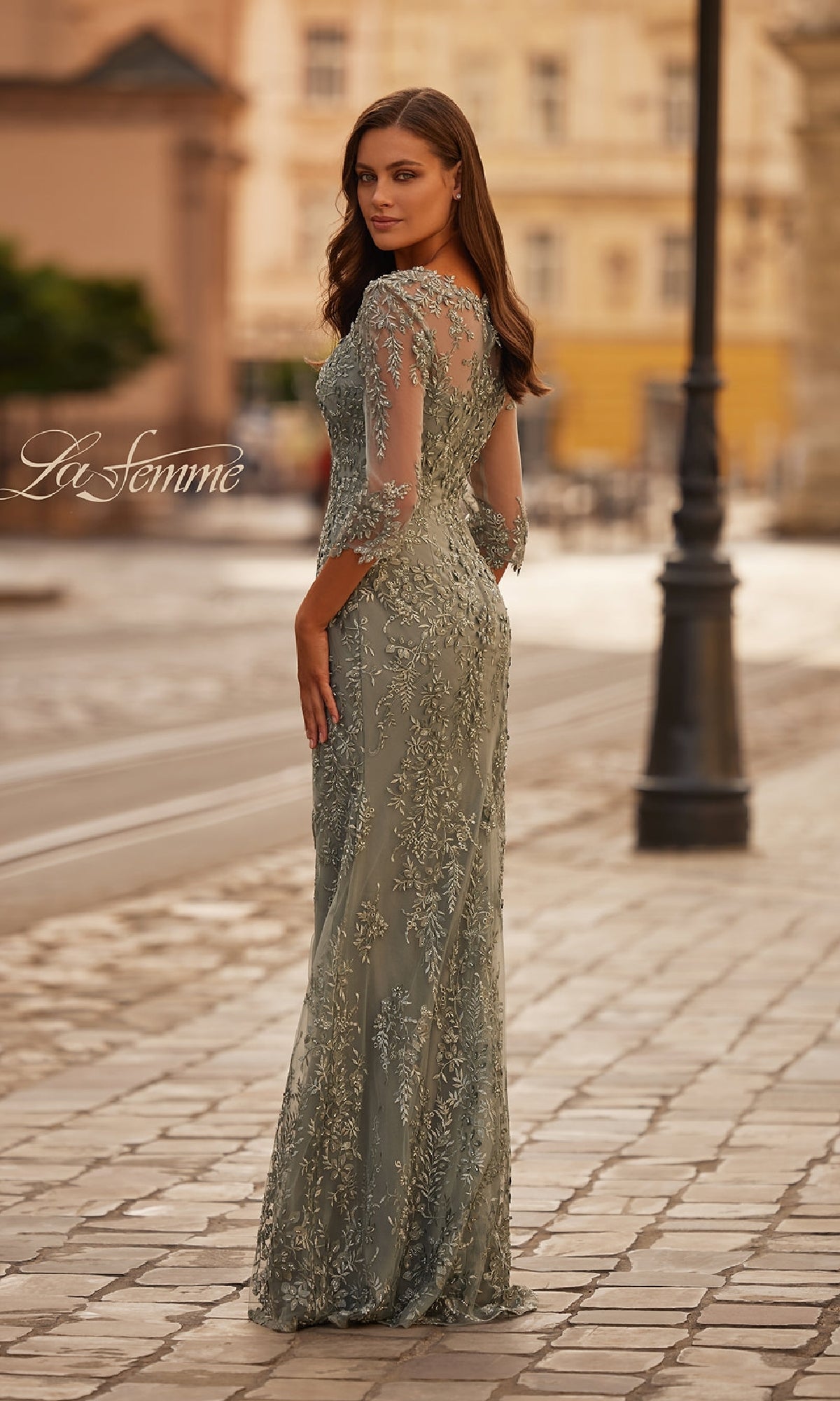 A woman stands on a cobblestone street in the La Femme 31909, a sage green lace long formal dress with sheer three-quarter sleeves and floral embroidery, looking back over her shoulder.